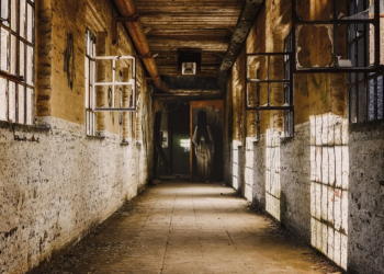 Dim abandoned corridor with broken windows and a painted silhouette of a robed figure at the far end