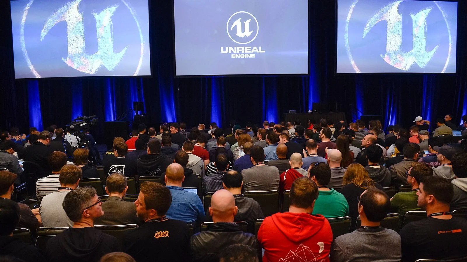 Audience at a conference watching the Unreal Engine logo displayed on three large screens