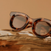 Round tortoiseshell sunglasses resting on driftwood against an amber background