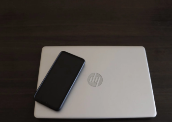 Closed silver HP laptop with round chrome logo on a dark desk, black phone resting on top