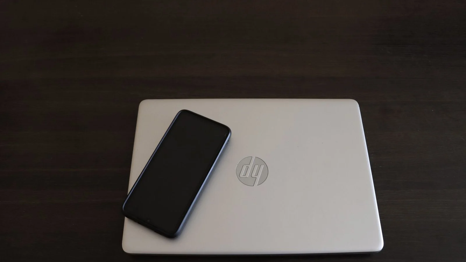 Closed silver HP laptop with round chrome logo on a dark desk, black phone resting on top