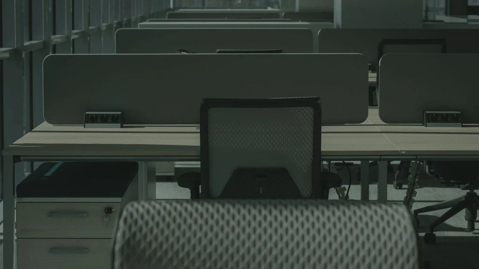 Empty office cubicles with unoccupied desk chairs and dividers in dim, greenish light
