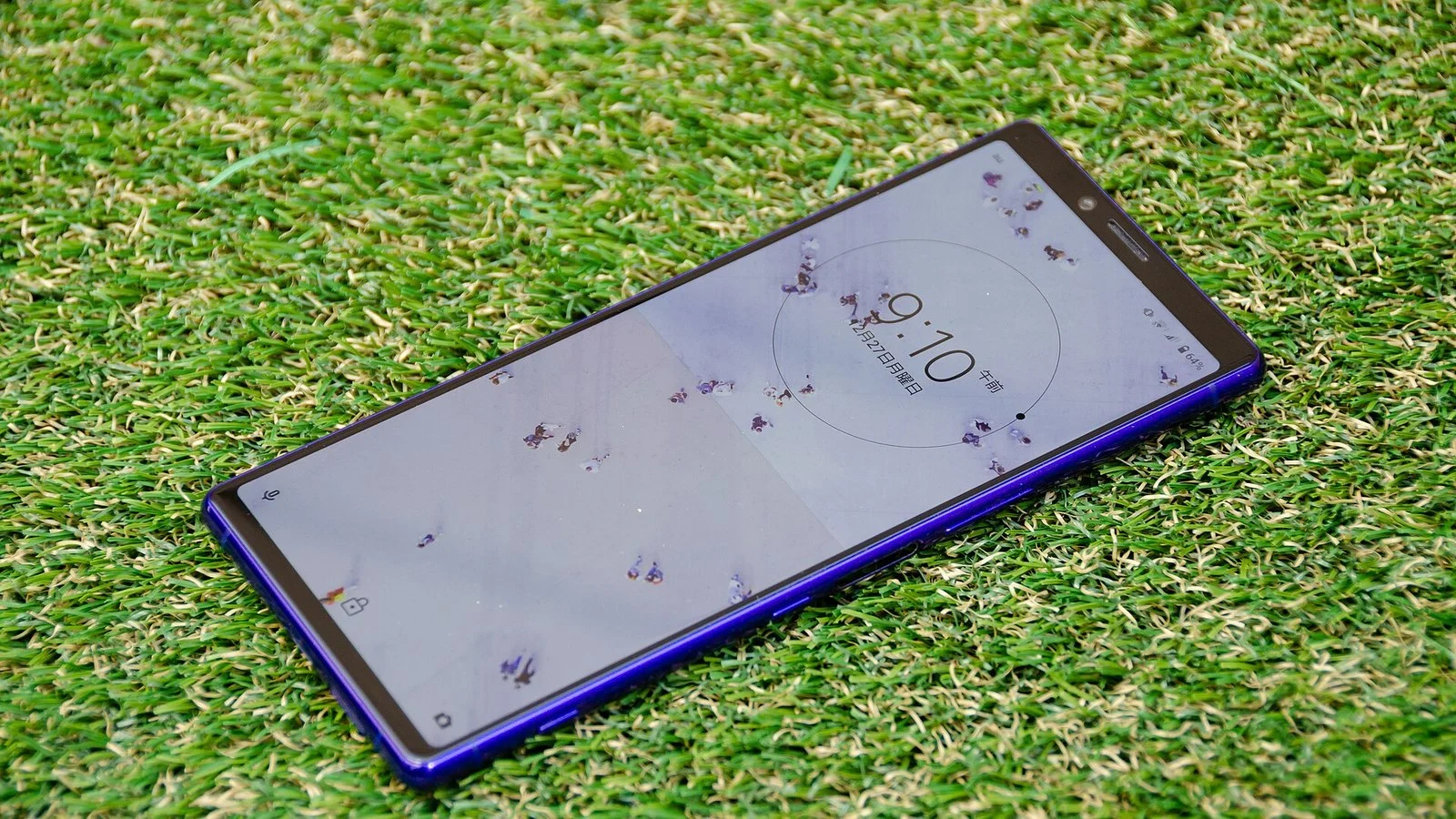 Blue Sony Xperia smartphone lying face-up on artificial grass showing a lock screen clock