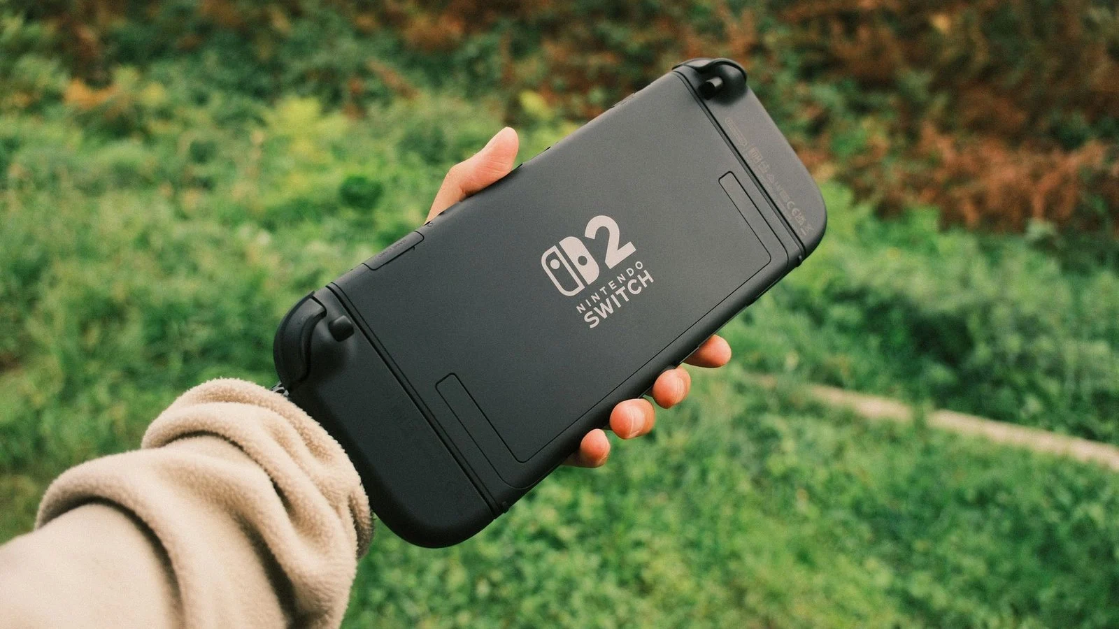 Hand holding the back of a black Nintendo Switch 2 console outdoors against greenery