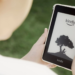 Person holding a white-bezel Kindle e-reader showing a tree-silhouette screensaver outdoors